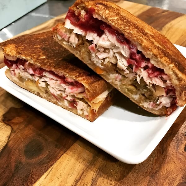 A grilled sandwich, cut in half, revealing turkey, cranberry sauce, and stuffing on a white plate.