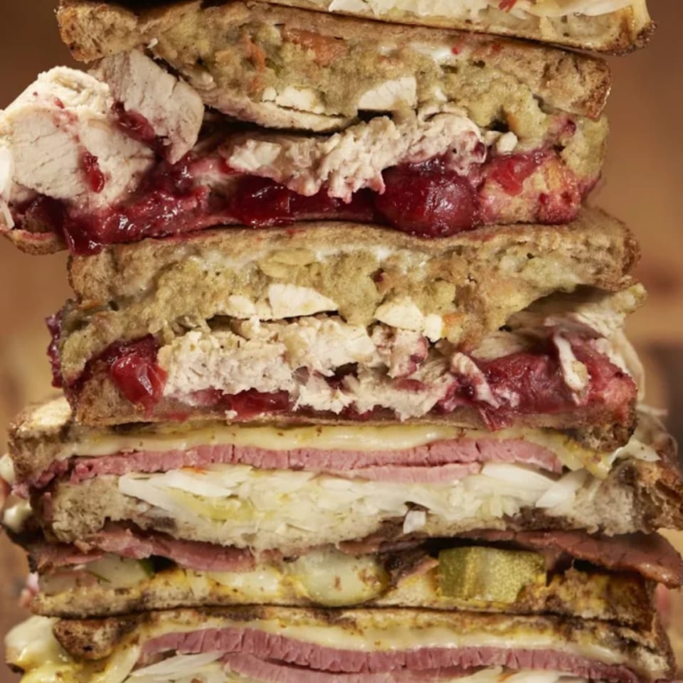 A towering stack of deli sandwiches, including turkey with cranberry, pastrami, and sauerkraut.